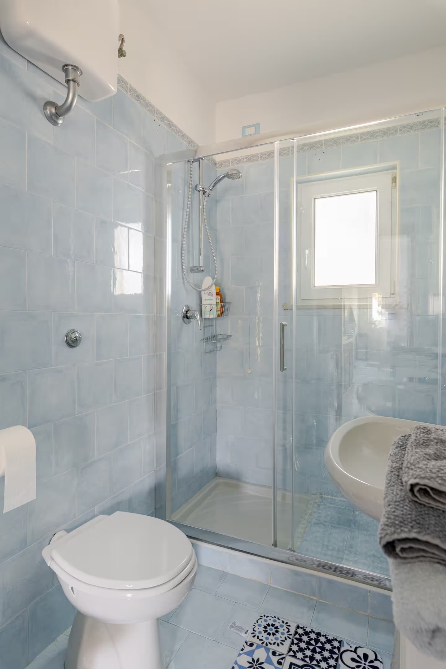 Bagno con piastrelle azzurre, box doccia in vetro, wc bianco e lavabo con asciugamani.