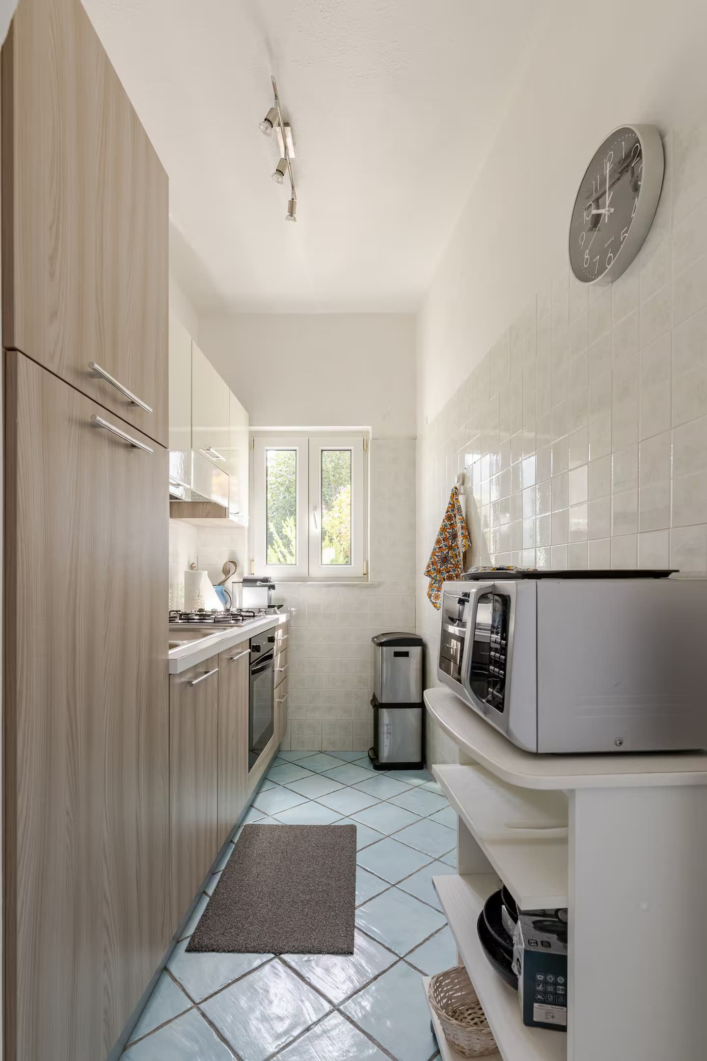 Cucina stretta con mobili in legno chiaro, pavimento azzurro e una finestra in fondo.