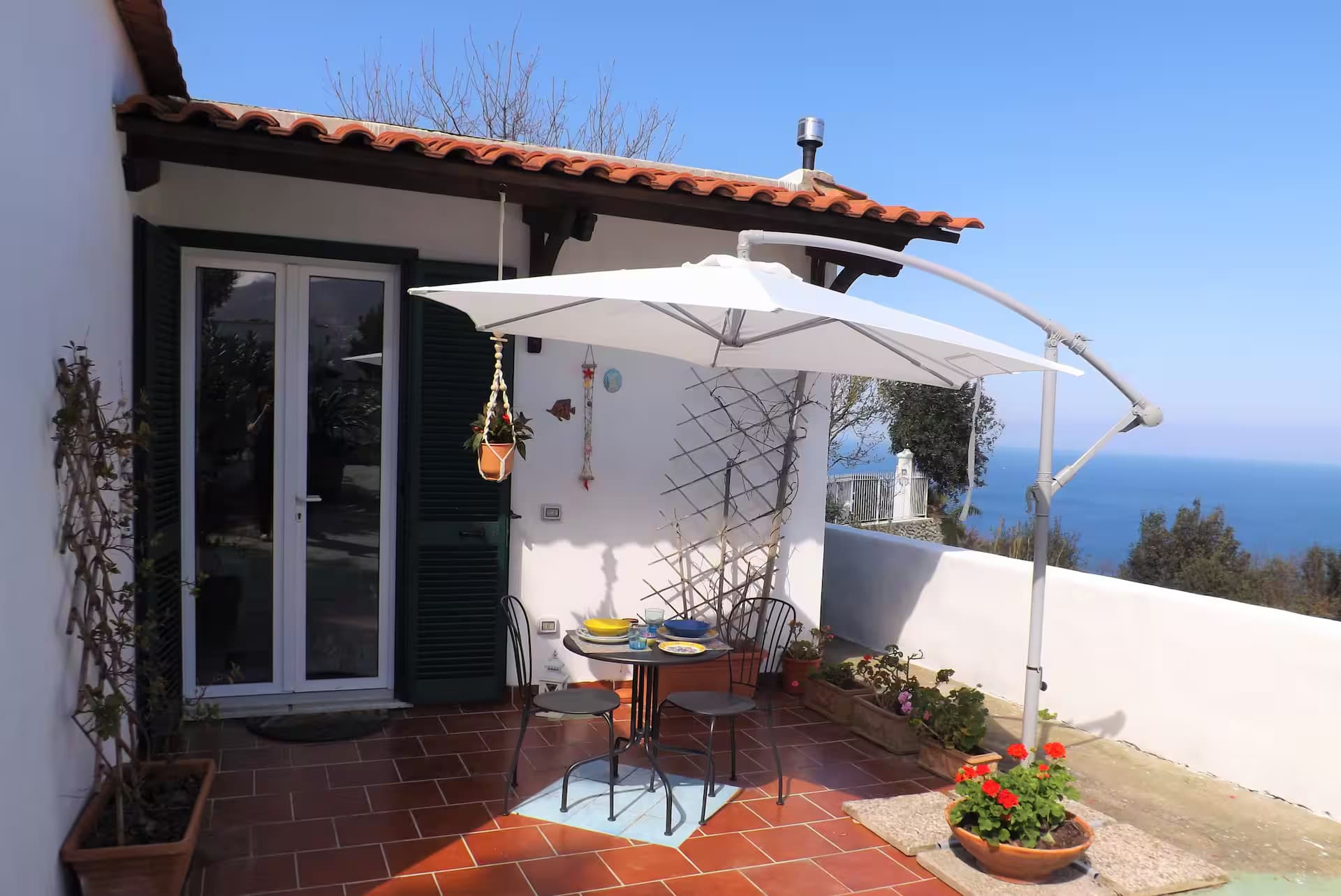 Terrazza soleggiata con ombrellone bianco, tavolo da pranzo e splendida vista sul mare blu.