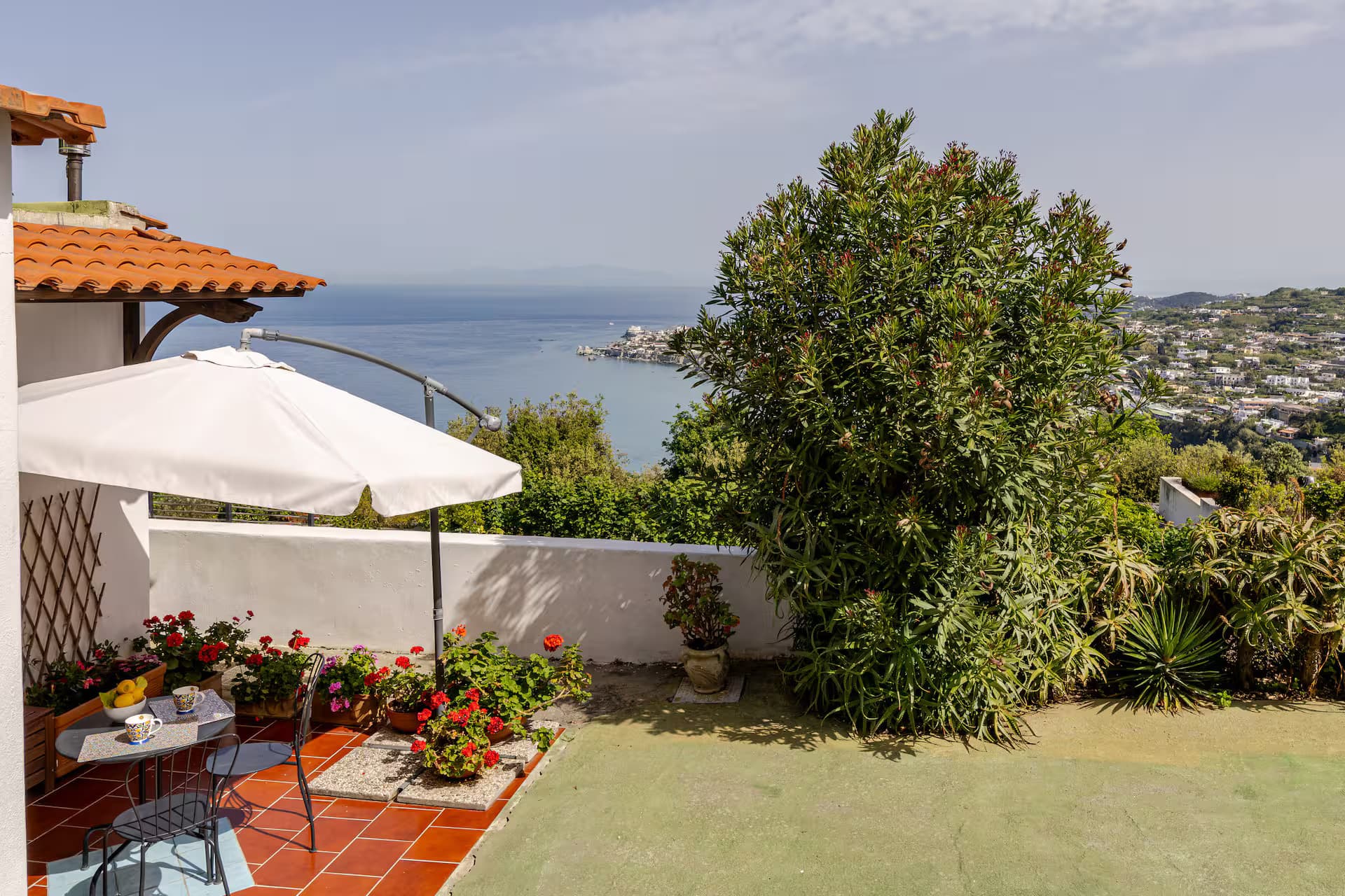 Terrazza con ombrellone bianco, fiori rossi e vista panoramica sul mare e sulla costa.