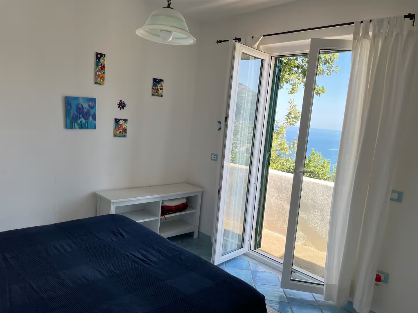 Camera da letto con copriletto blu e porte finestre aperte con vista sul mare.