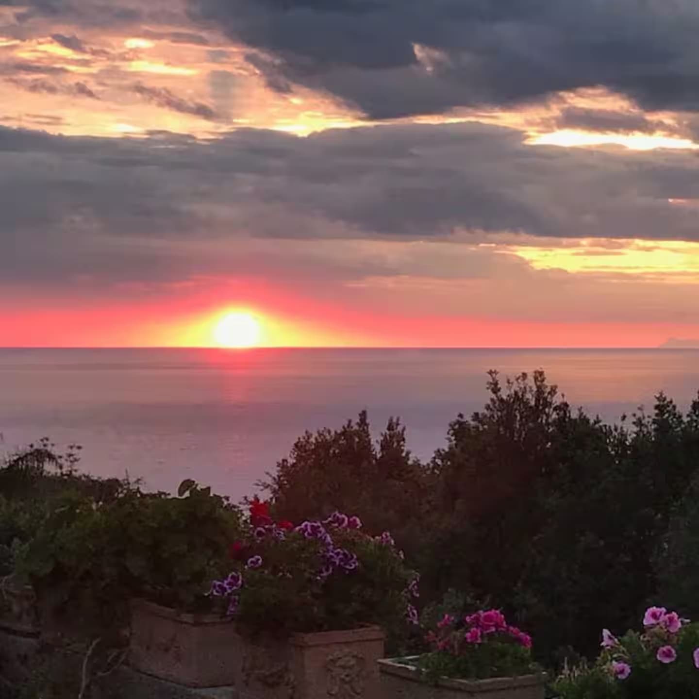 Tramonto rosso sul mare con nuvole scure e vasi di fiori in primo piano.